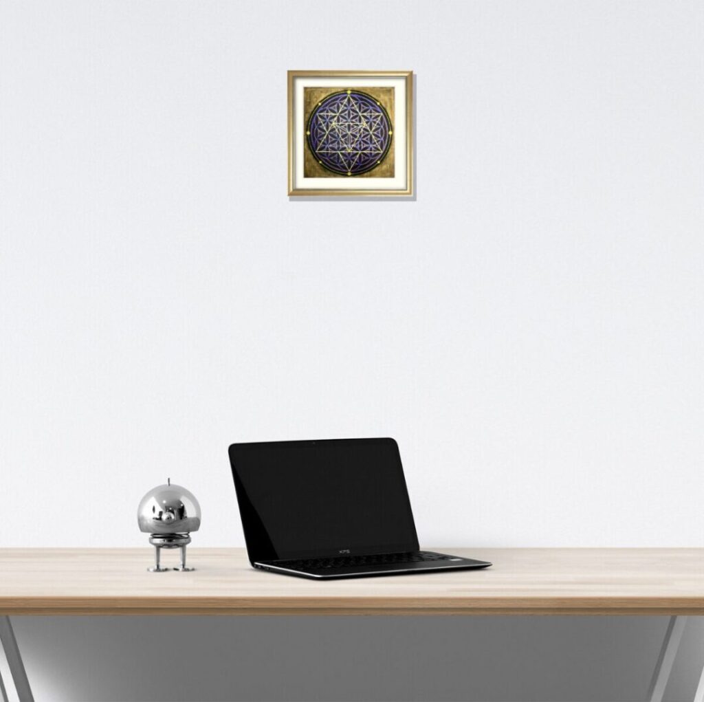 ベージュのデスクとパソコンのある空間。白い壁に飾られたマカバアート「宇宙」が、シャンパンゴールド